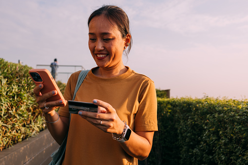 A smiling Asian woman shopping online in the park, typing in payment information from her credit card into her mobile phone.