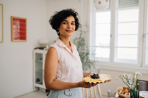 A happy Latin American woman standing proudly by the dining table, holding a cheese board.