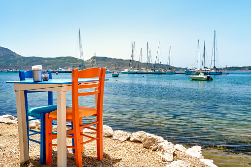 Colorful chairs and tables of Greek tavern by waterfront at sunny day. Vacations in Greece, summer, restaurant, outdoor dining, port, marina, yachts and boats, Mediterranean relax, Cyclades and Aegean islands. Selective focus, blurred background.