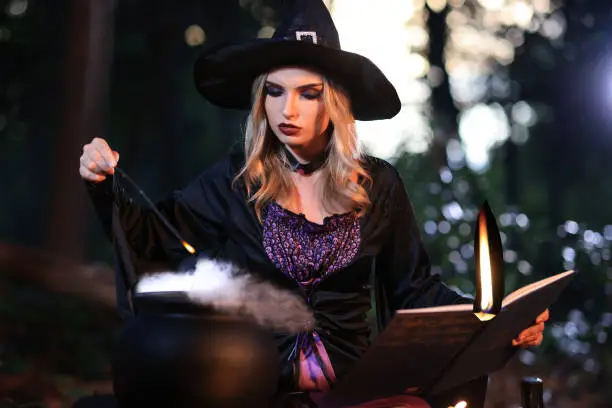 Photo of The fantasy woman witch conjures, holds a magic wand and a magic book in her hands, and reads spell white magic smoke from boiling vat. Halloween theme. Witch performs ceremony on Halloween day