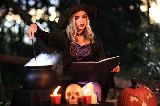 The fantasy woman witch conjures, holds a magic wand and a magic book in her hands, and reads spell white magic smoke from boiling vat. Halloween theme. Witch performs ceremony on Halloween day