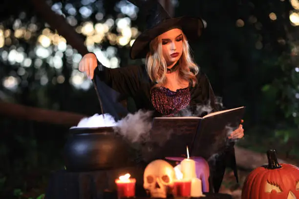 Photo of The fantasy woman witch conjures, holds a magic wand and a magic book in her hands, and reads spell white magic smoke from boiling vat. Halloween theme. Witch performs ceremony on Halloween day