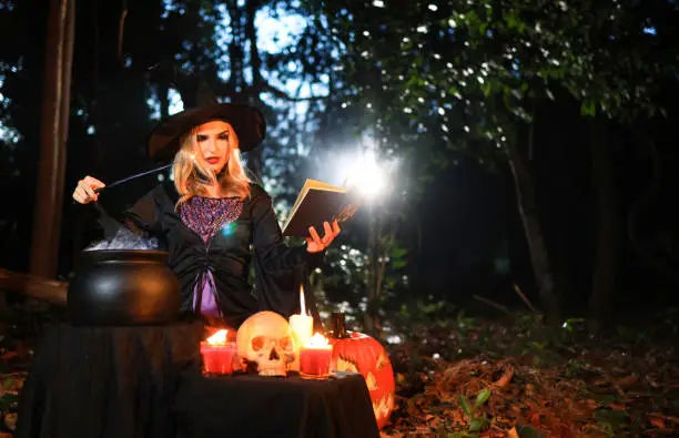 Photo of The fantasy woman witch conjures, holds a magic wand and a magic book in her hands, and reads spell white magic smoke from boiling vat. Halloween theme. Witch performs ceremony on Halloween day