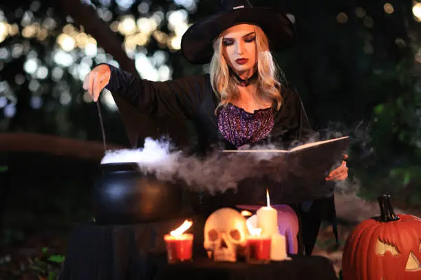 Photo of The fantasy woman witch conjures, holds a magic wand and a magic book in her hands, and reads spell white magic smoke from boiling vat. Halloween theme. Witch performs ceremony on Halloween day