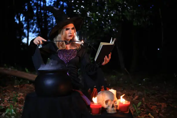 Photo of The fantasy woman witch conjures, holds a magic wand and a magic book in her hands, and reads spell white magic smoke from boiling vat. Halloween theme. Witch performs ceremony on Halloween day