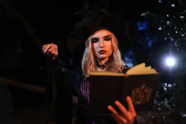 Photo of The fantasy woman witch conjures, holds a magic wand and a magic book in her hands, and reads spell white magic smoke from boiling vat. Halloween theme. Witch performs ceremony on Halloween day