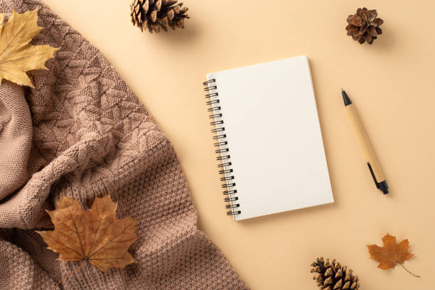 istock Fall-themed office setup: Overhead shot of warm knitted blanket, notepad, pen, maple leaves and pine cones. Soft beige backdrop, perfect for messaging 1657611635