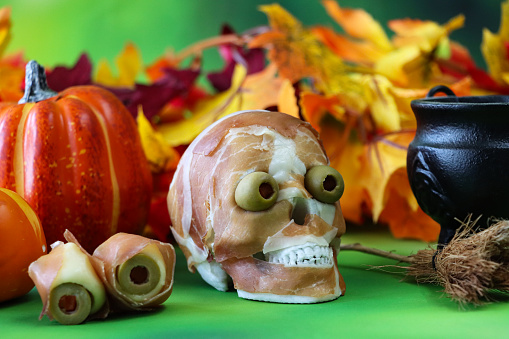 Stock photo showing close-up view of a Halloween scene with a Prosciutto di Parma skull with red pepper stuffed green olive eyeballs besides a witch's black cauldron, with artificial autumnal fallen leaves against a mottled green background.