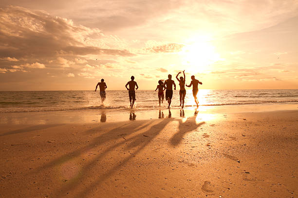 Group of multi ethnic friends having fun at the beach Six young adults are running on the beach towards the water.  The sky has a number of clouds, with the majority of the clouds on the left.  The sun is low in the sky on the right half of the sky and is slightly covered with clouds. beach party stock pictures, royalty-free photos & images