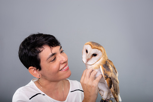 A woman holding and touching a baby owl. Wild animal. Studio portrait against gray background.