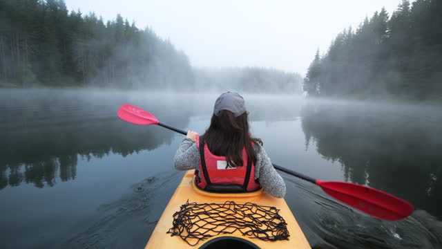 Rear view of woman kayaking on a mountain foggy lake in the morning.