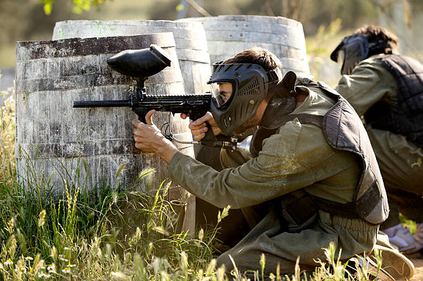 Paintball player Protective sport player in uniform and mask Aiming gun, behind a barrel paintballing stock pictures, royalty-free photos & images