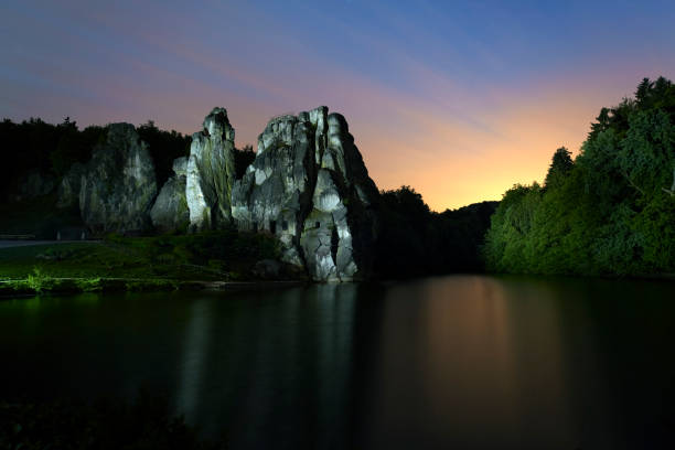Rock behind lake at dusk with majestic sky. Rock and forest illuminated by myself with special flashlight. Long exposure with blurred motion and very little noise on water surface of lake in the foreground. 