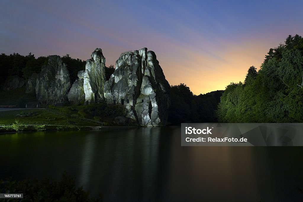 Illuminated rock at dusk - Externsteine in North Rhine Westfalia Rock behind lake at dusk with majestic sky. Rock and forest illuminated by myself with special flashlight. Long exposure with blurred motion and very little noise on water surface of lake in the foreground.  Externsteine Stock Photo