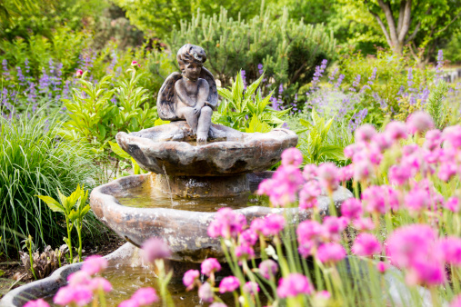 A winged angel sits on a bronze-colored plaster fountain in a garden.