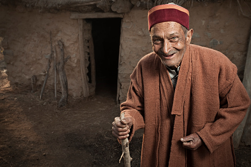 Real people from rural India: Happy Senior man looking at camera and standing near adobe cattle house by holding stick. He is wearing traditional dress of Himachal Pradesh, India.