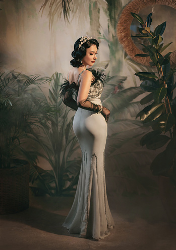 Sexy back, elegant woman in twenties old style mermaid silhouette silver retro long evening dress black ostrich feather boa cold wave hairstyle pearl crown. vintage art photo. Roar 1920s fashion model