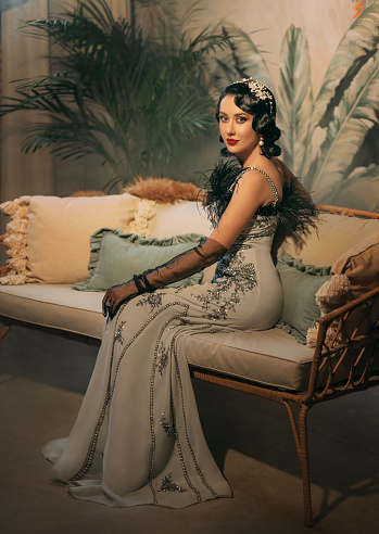 1920s style Happy sexy flapper woman sitting on sofa, elegant gray silver dress black ostrich feather. Roar fantasy girl beauty face. classic room. white pearl beads headband, cold wave hairstyle 20s.