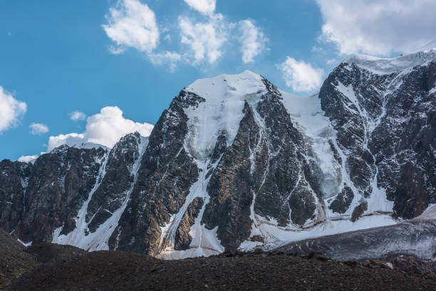 Atmospheric landscape with large snow mountain range under clouds in blue sky. Awesome high snowy mountains in changeable weather. Dramatic scenery with snow-covered mountain wall under cloudy sky.