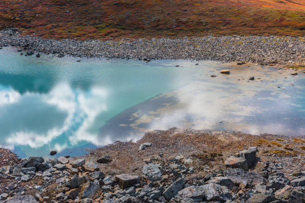 istock Abstract nature background with sunlight through clouds reflected in turquoise mountain lake mirror. Beautiful pure alpine lake with cloudy sky reflection among vivid autumn colors in high mountains. 1657991544