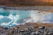 Abstract nature background with sunlight through clouds reflected in turquoise mountain lake mirror. Beautiful pure alpine lake with cloudy sky reflection among vivid autumn colors in high mountains.