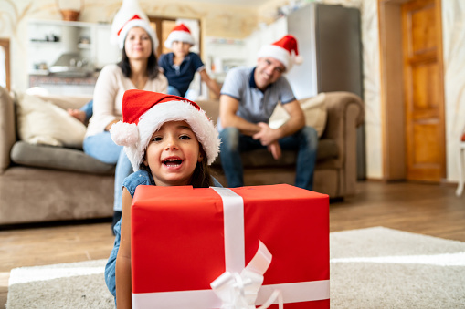 A girl is looking at the camera over the top of a large Christmas present wrapped in red paper, her family wearing santa hats are behind her sitting on a sofa