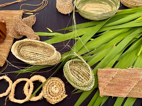 Horizontal high angle closeup photo of various items hand woven from dried Australian native plants and a bunch of green bullrush leaves on a black cloth background.
