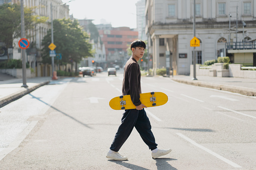 An Asian Chinese young man carrying skateboard walking across the street in a city