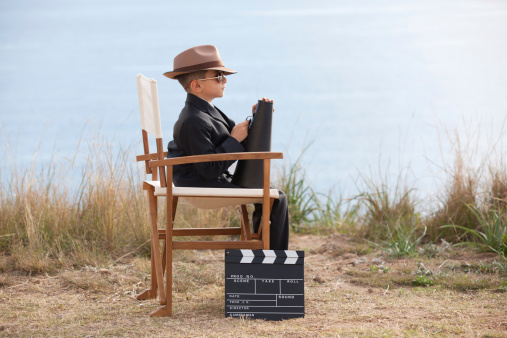 Little Boy Holding Megaphone And Sitting On Director's Chair in outdoor film set,full length director's chair and film slate in frame