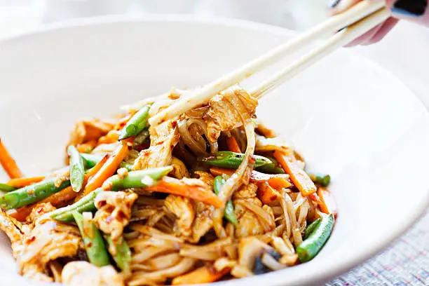 A woman's hand uses chopsticks to lift a portion of  a Thai dish of chicken, stir fried with green beans, julienne carrots. chilli bits and noodles, from a white china bowl.