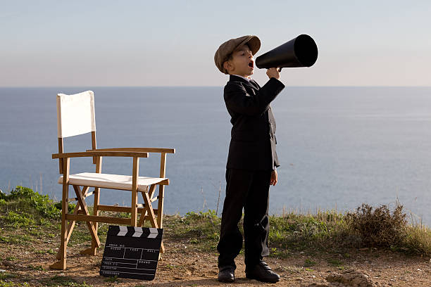 Little Film Director Shouting With Megaphone In Outdoor Set Photo of little boy in full length standing and shouting on old fashioned megaphone in outdoor.A director chair and film slate is seen just close to him.He is wearing a flat cap and a black suit.Sea is seen on the background.The image was shot in daylight with a full frame DSLR camera. directing photos stock pictures, royalty-free photos & images