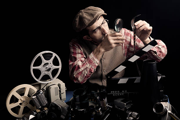 Adult Man Holding Cinema Film And Checking With Magnifying Glass Adult Man Holding Cinema Film And Checking With Magnifying Glass for editing.There are large amount of cinema objects placed on desk in front of model.A 8 mm film projector, film slate,video cameras, microphone, film reels etc are seen.Model is wearing a flat newsboy cap, waist and a plaid red shirt.Photo was shot with a full frame DSLR camera in studio. editing equipment stock pictures, royalty-free photos & images