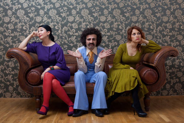 Sullen two women and a men sitting on sofa.They are sitting on a leather brown sofa in front of wallpaper background.The woman on the left is wearing a purple dress and red socks while men is sitting in the middle and fearing 1970s style waist and pants.The other woman is sitting on the right side of frame  is wearing a green dress.Horizontal, studio shot.
