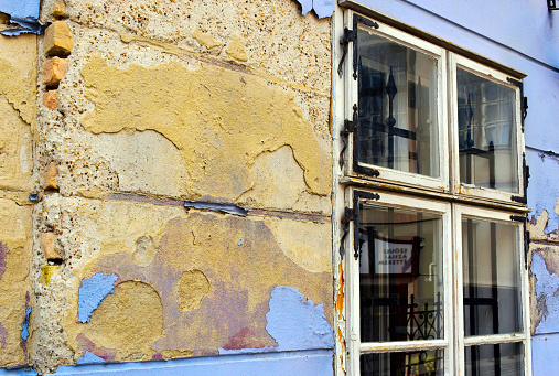 grunge deteriorating building exterior elevation stucco finish. exposed damaged, spalling brick wall due to moisture. building renovation concept. peeling yellow and blue paint. weathered wood window