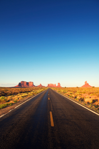 Wide open American road goes off into the horizon featuring jagged mesa formations of Monument Valley 
