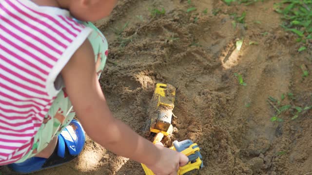 Asian boy plays and digs sand using a toy digger and dump truck.