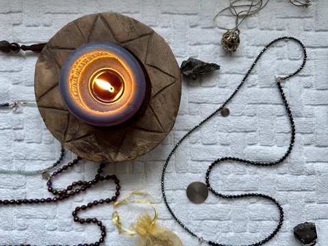 Top view of a burning scented candle on a round wooden carved holder placed on a white tablecloth surrounded by beaded jewellery