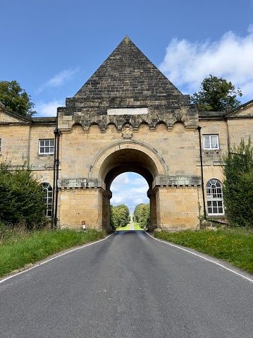 A portrait oriented photograph produced from outside the gatehouse of Castle Howard on the North Yorkshire Moors near York in the Yorkshire Dales, England. The long straight public highway can be seen and through the archway the 100 foot obelisk can be seen in the distance. The photograph was produced on a bright sunny day.