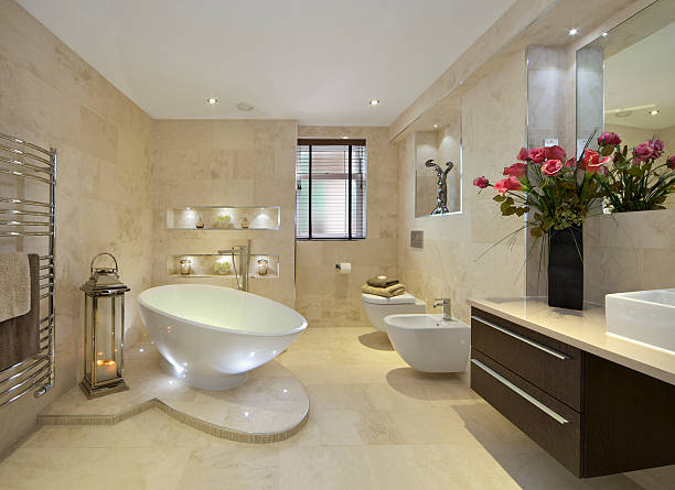 elegant bathroom with flowers a modern bathroom in an expensive new home with a tear-drop shaped bath (full of water) sitting on a marble plinth. A bidet and WC are located near the window. A lantern with lit candles sits next to a large towel rail whilst a large bunch of red lilies sit on the cabinet to the right of the picture. light brown bathroom stock pictures, royalty-free photos & images