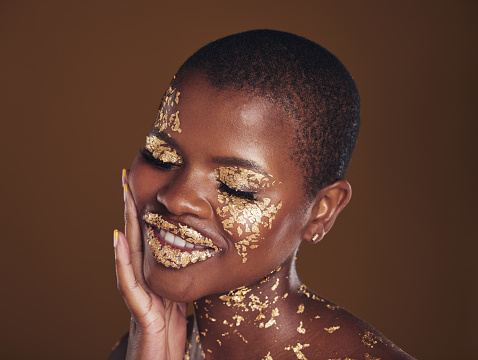 Black woman, relax and gold makeup with luxury dermatology and skincare mask with glitter. Sparkle, smile and beauty with happy glow and shine of cosmetics and model in a studio with facial art