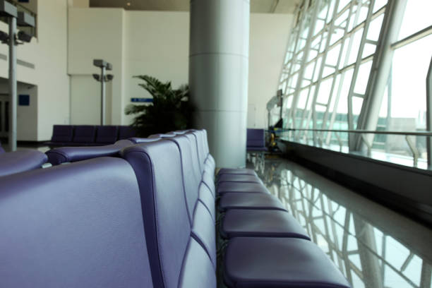 A quiet and modern airport waiting area with a row of empty black chairs positioned in front of large terminal windows. The soft natural light from outside enhances the peaceful and spacious atmosphere, offering a glimpse of the runway in the background. Ideal for travel, transportation, waiting, journey, or business-themed concepts.