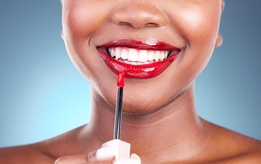 Woman, smile and red lipstick with brush and beauty, happy with makeup closeup isolated on blue background. Model, bold and cosmetic product on lips, mouth with skin and teeth with shine in studio