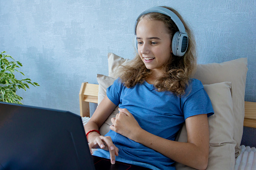 Cute teenage girl in wireless headphones listening to music on her laptop on the bed at home.