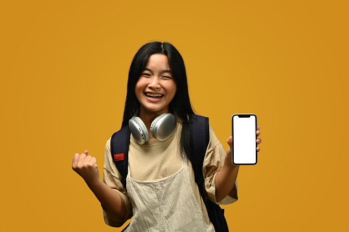 Teenager schoolgirl with backpack showing smartphone with blank screen on pink background.