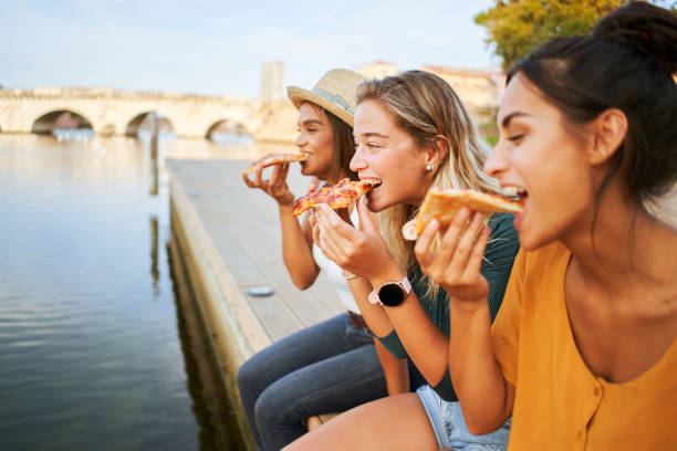 Three beautiful girls eat pizza on the lakeside. Group of women enjoy free time together sightseeing in a roman city in europe. stock photo