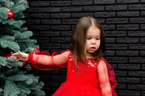 Little girl in red dress smiling by Christmas tree. Little beautiful girl in a red evening dress the Christmas tree. Girl in red in Christmas decorations. Christmas concept. New Year. Baby near a Christmas tree. High quality photo.