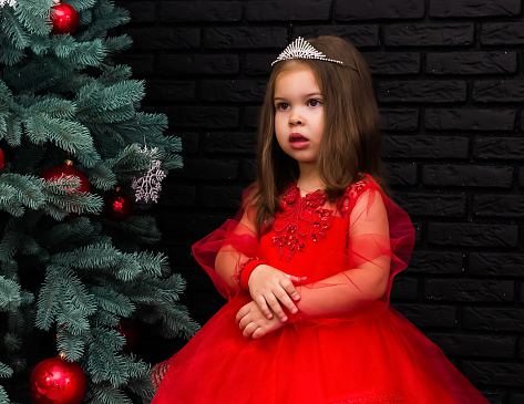 Little girl in red dress smiling by Christmas tree. Little beautiful girl in a red evening dress the Christmas tree. Girl in red in Christmas decorations. Christmas concept. New Year. Baby near a Christmas tree. High quality photo.