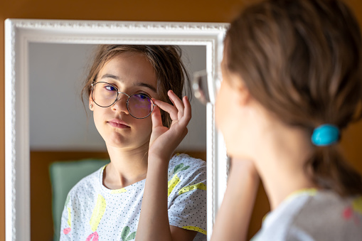 Sleepy teen girl in glasses looks at herself in the mirror in the morning, the concept of self-acceptance.