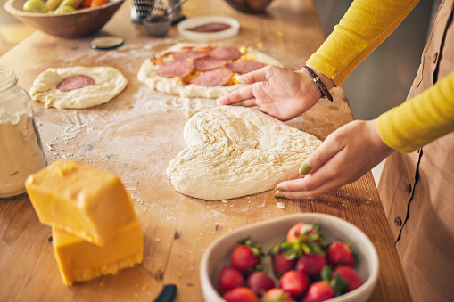 Pizza, heart with dough and hands on wooden table, person cooking with flour and cheese, fruit and food at home. Nutrition, meal and strawberry in kitchen, baking with ingredients and love sign