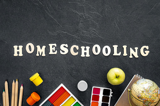 Homeschooling word made of wooden letters and colorful school supplies and stationery on a dark textured board for your design, top view.